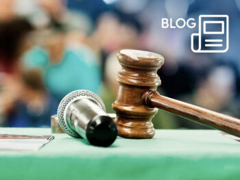 Close-up of a wooden gavel and a microphone on a green table, with a blurred crowd in the background for an auction room post.
